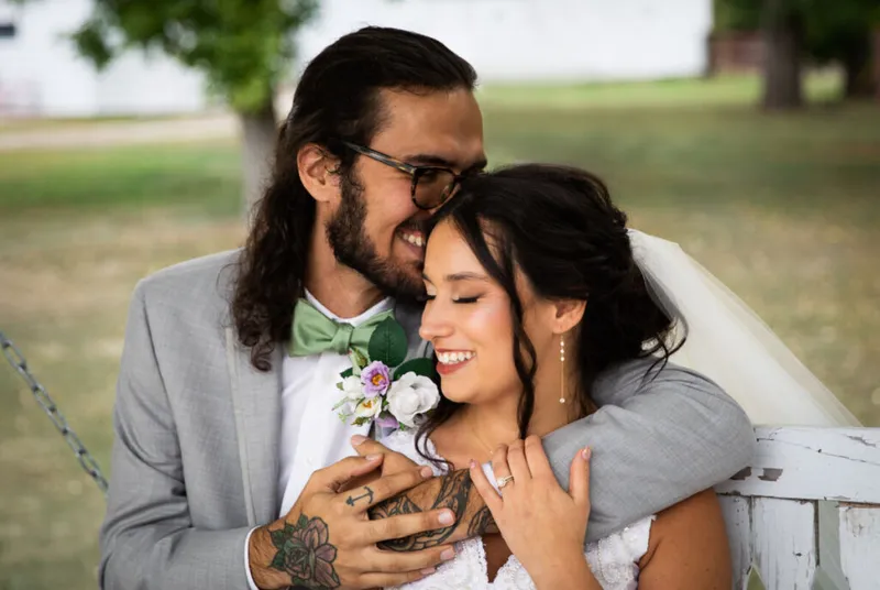 Smiling bride in veil and groom in grey suit with green bow tie embrace on a white swing outdoors