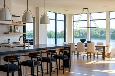 Modern kitchen island with dark countertop and bar stools overlooking a sunlit lake through floor-to-ceiling windows