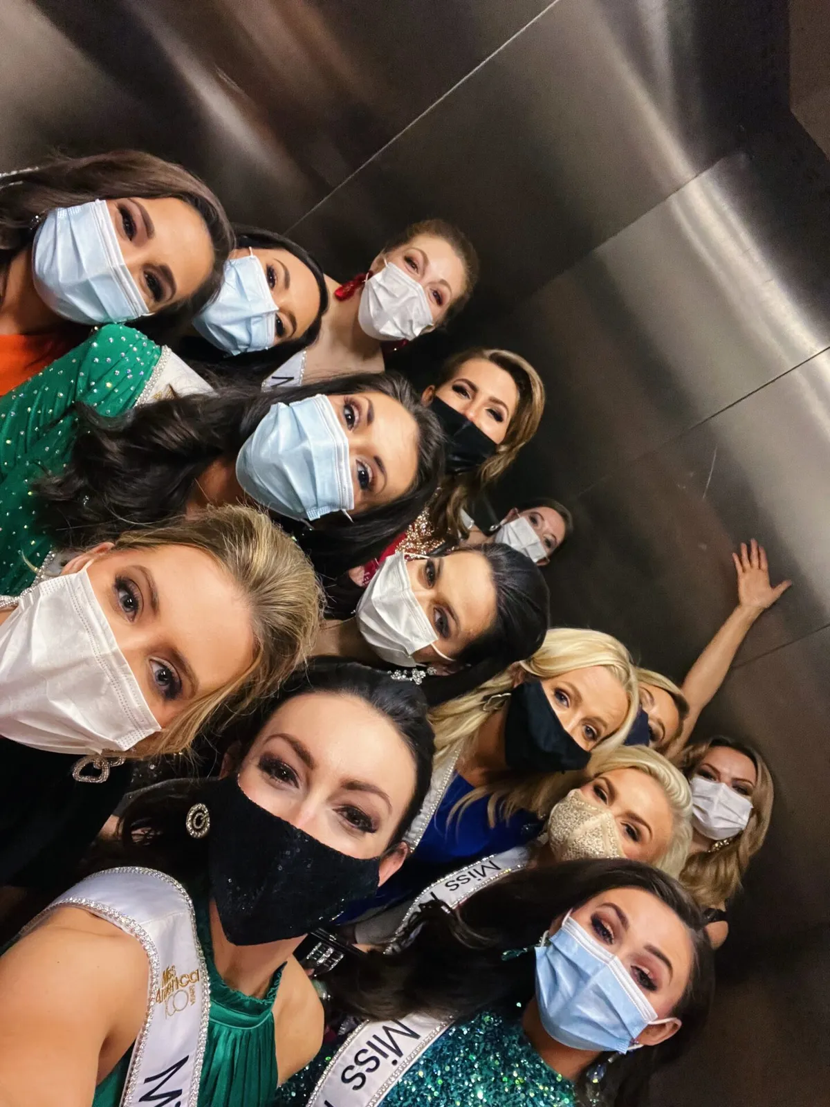 Group of masked pageant contestants wearing sashes crowd together for a selfie in an elevator