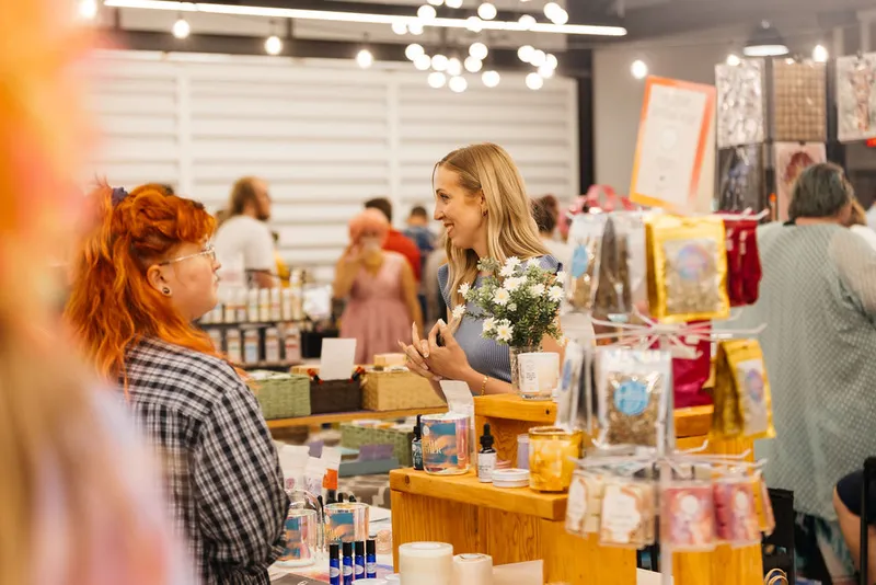 Two women chat across a vendor table stocked with candles and packaged goods at an indoor market