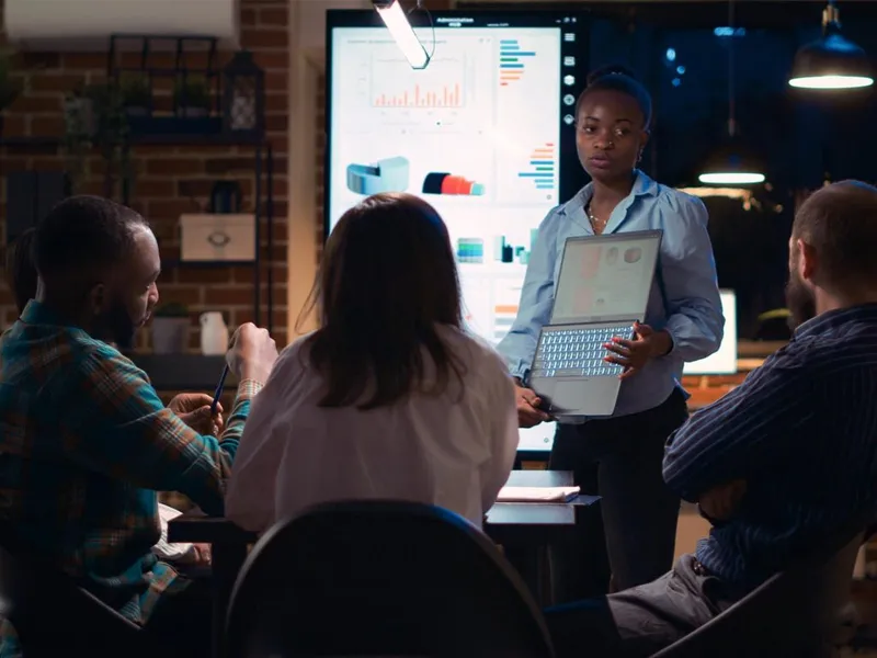 Woman holds laptop while presenting data charts to three seated colleagues in a dim office