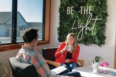 Two people chat at a table beneath a green moss wall with "Be The Light" sign
