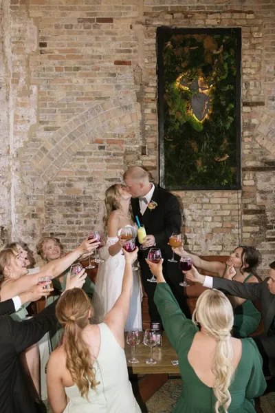 Bride and groom kiss as wedding guests raise wine glasses in a toast inside a brick-walled venue