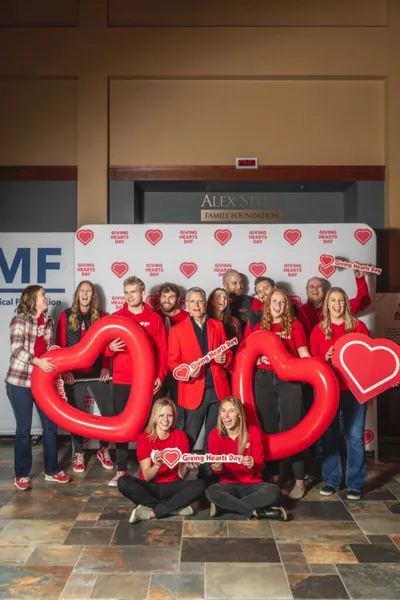 Group posing with large red inflatable hearts and signs in front of a Giving Hearts Day step-and-repeat banner