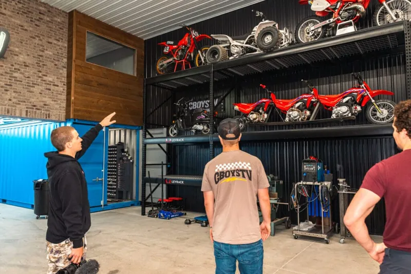 Three men look up at red dirt bikes stored on elevated racks inside a garage