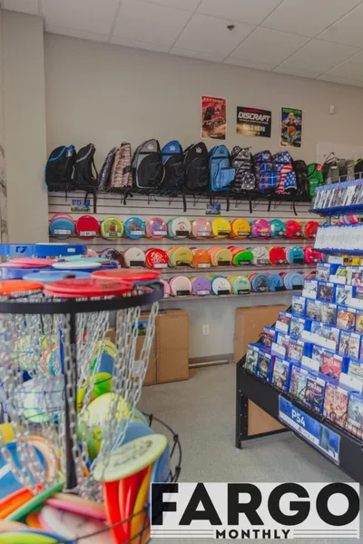 Disc golf store interior with colorful discs on shelves, backpacks on wall, and a basket in the foreground