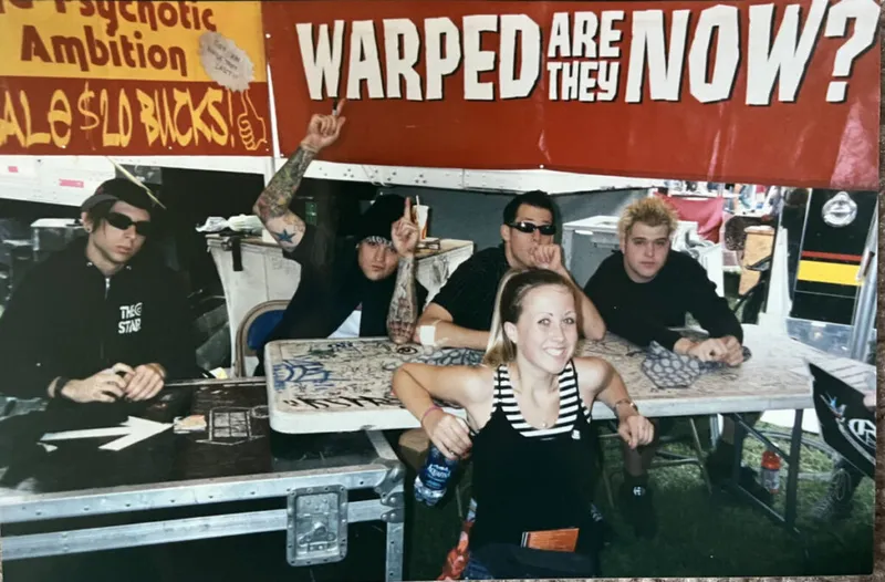 Group of young people at a Warped Tour merch table under a red 'Warped Are They Now?' banner
