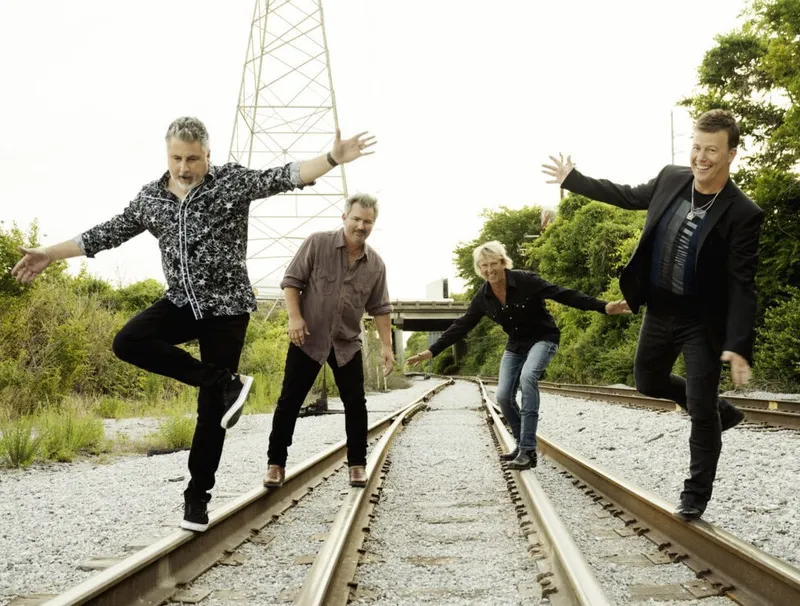 Four adults playfully balancing on railroad tracks surrounded by green foliage