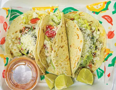 Three flour tortilla tacos topped with lettuce, tomato, and crumbled cheese, served with lime wedges and salsa