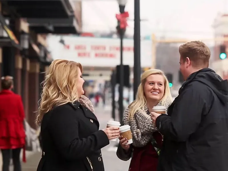 Three people chatting on a city sidewalk in winter coats, holding takeaway coffee cups