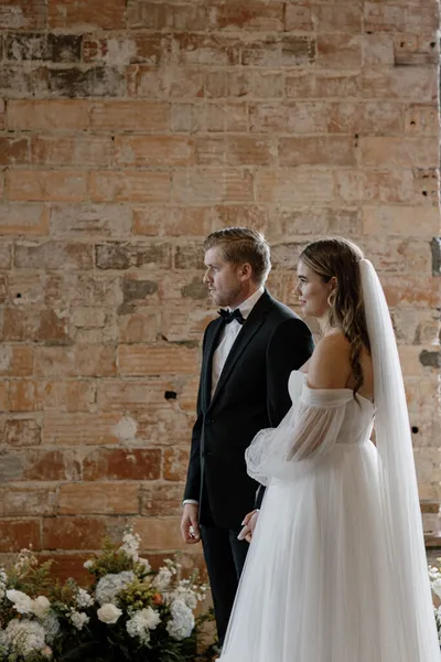 Bride in off-shoulder gown and veil stands beside groom in black tuxedo before a rustic brick wall