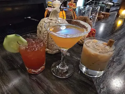 Three cocktails on a dark bar top, including a red drink with lime, a sugared-rim martini, and a cream-colored drink with a