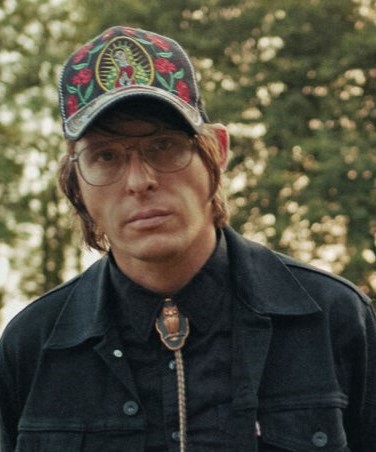 Man in embroidered floral cap, aviator glasses, and bolo tie outdoors