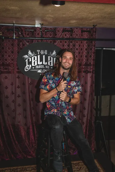 Long-haired man in a floral shirt holds a microphone on stage at The Cellar in Fargo, ND