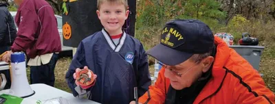 A smiling boy holds a small item beside an older man in a U.S. Navy cap at an outdoor fall festival booth