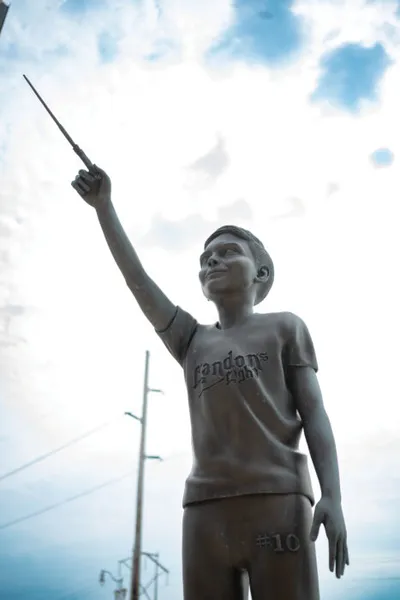 Bronze statue of a boy raising a wand overhead, wearing a shirt reading 'Landons' and #10