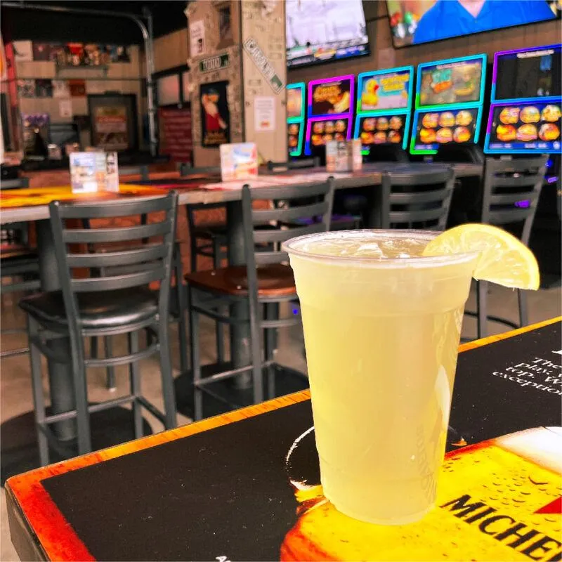Lemonade in a plastic cup with a lemon wedge on a bar table, arcade machines glowing in the background