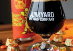 Dark beer in a stemmed glass beside Junkyard Brewing Company signage and colorful cans