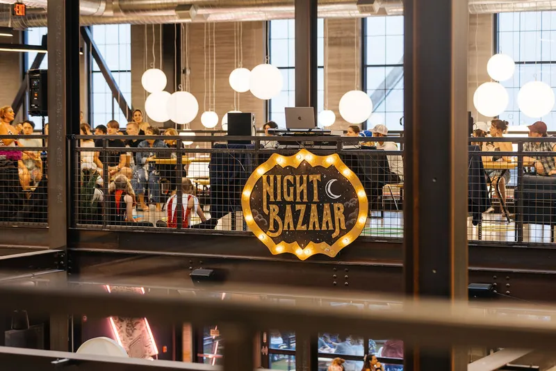 Illuminated Night Bazaar marquee sign on a mezzanine railing surrounded by globe pendant lights and a crowd