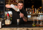 Bartender pouring from a bottle into a metal shaker at a dimly lit bar