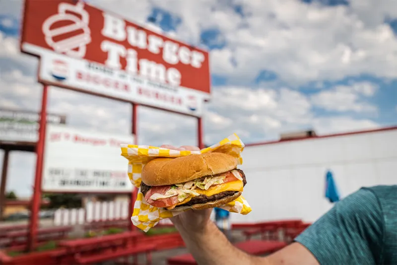 Hand holding a cheeseburger in checkered paper in front of a Burger Time sign