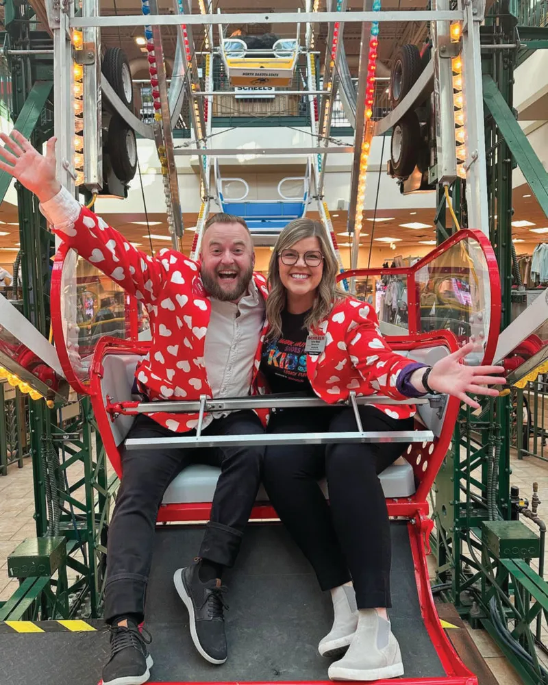 Two smiling people in matching red heart-print jackets ride an indoor Ferris wheel gondola with arms outstretched