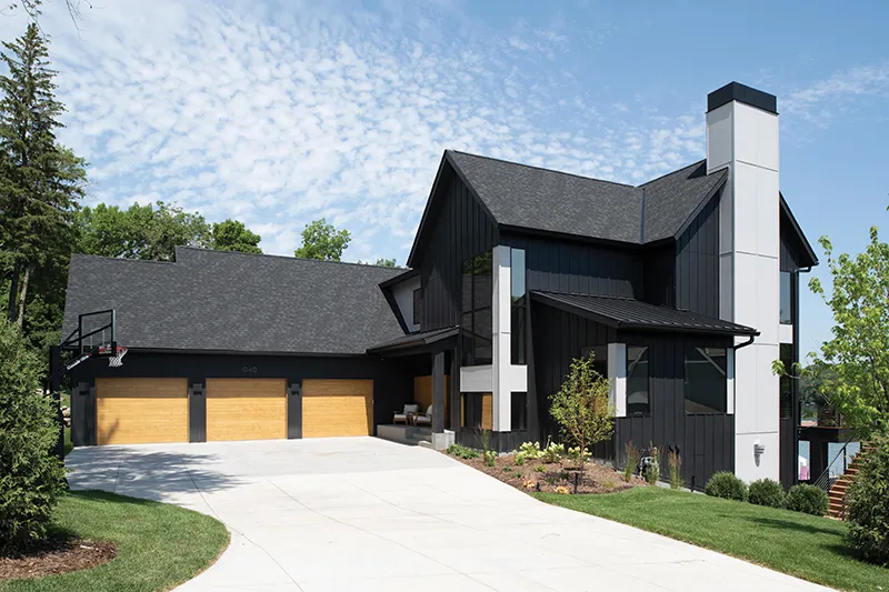 Modern black board-and-batten home with wood garage doors, white chimney, and concrete driveway