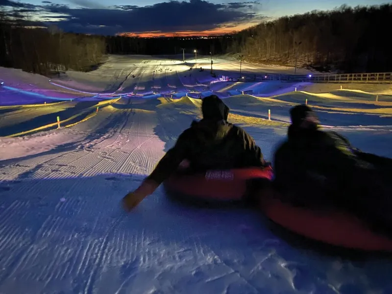 Two people on red snow tubes descend a lit tubing hill at dusk