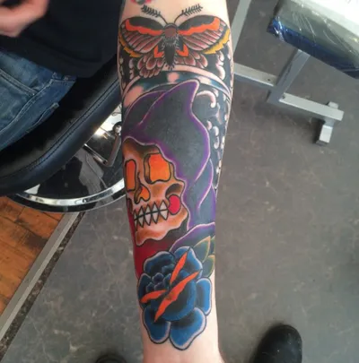 Forearm tattoo sleeve featuring a grim reaper skull, blue rose, and moth in bold traditional style