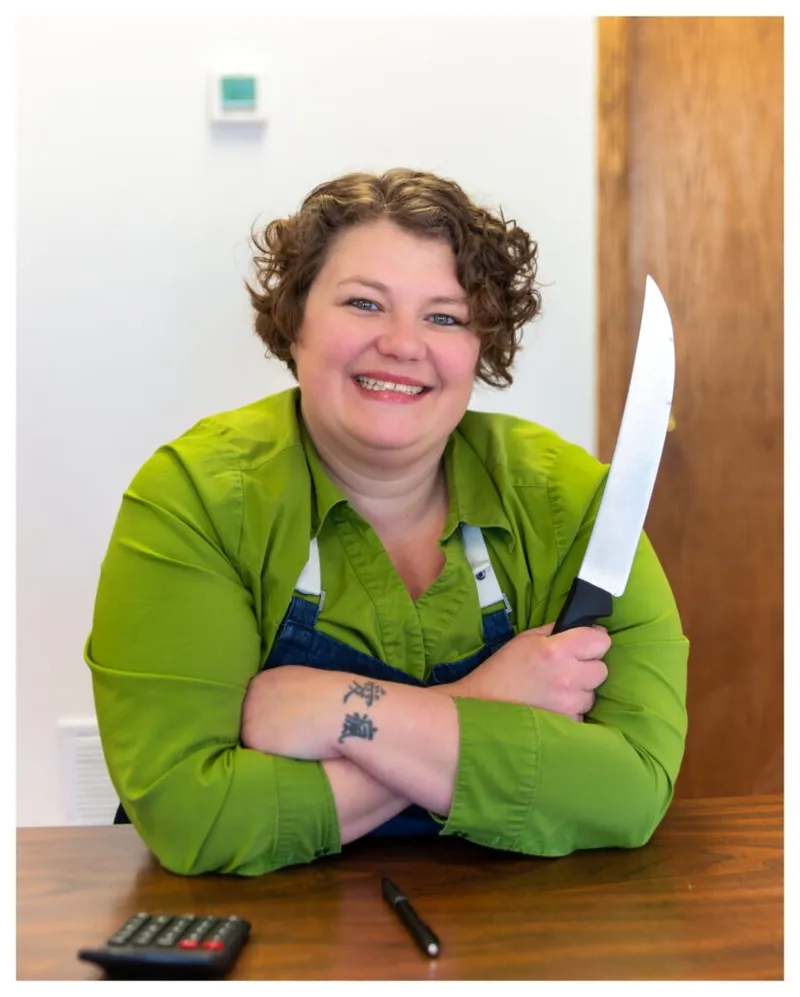 Smiling woman in green shirt and denim overalls holding a large chef's knife, arms crossed at a wooden table