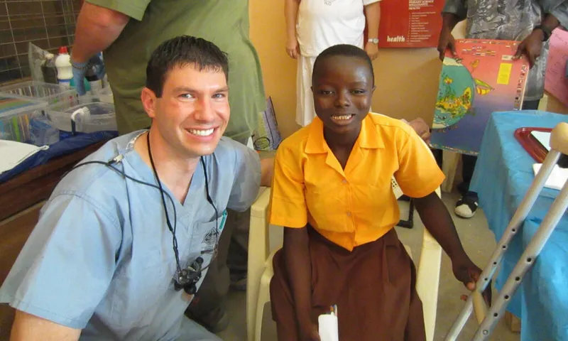 Smiling dentist in blue scrubs crouches beside a young child seated in a clinic chair