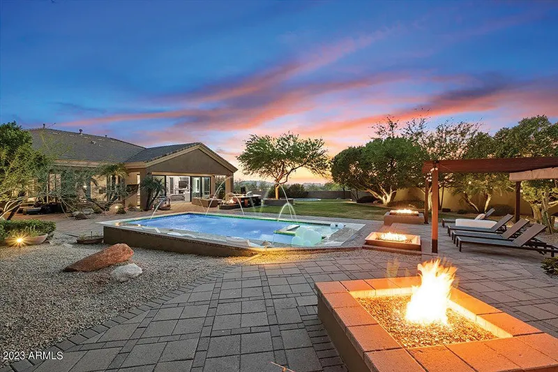 Backyard pool with fountains, two lit fire pits, lounge chairs, and a pergola at dusk