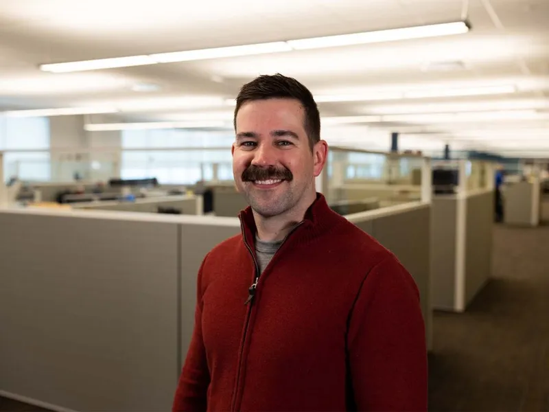 Smiling man with a mustache in a red zip-up jacket standing in an open office with cubicles