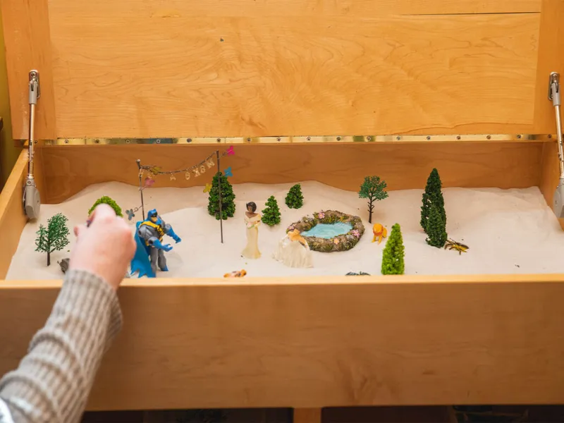 A hand arranges miniature figurines and trees in a sand tray therapy box