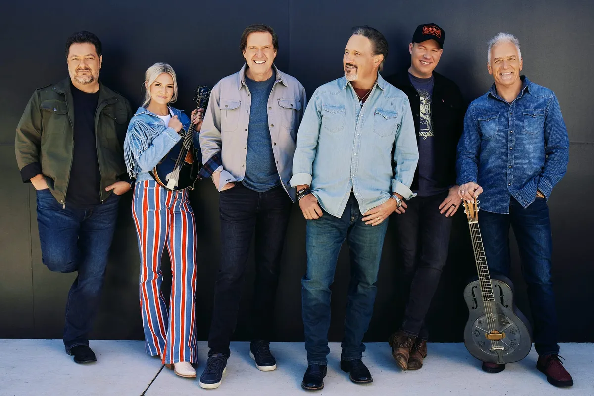 Six smiling musicians pose against a dark wall, one holding a mandolin and another a resonator guitar