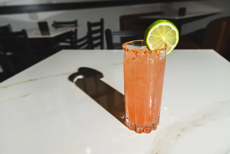 Pink cocktail in a ribbed highball glass with a chili-salt rim and lime wheel on a marble bar