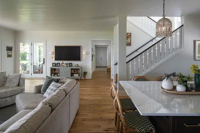 Open-plan living room with gray sofa and wall-mounted TV beside a white staircase and marble kitchen island