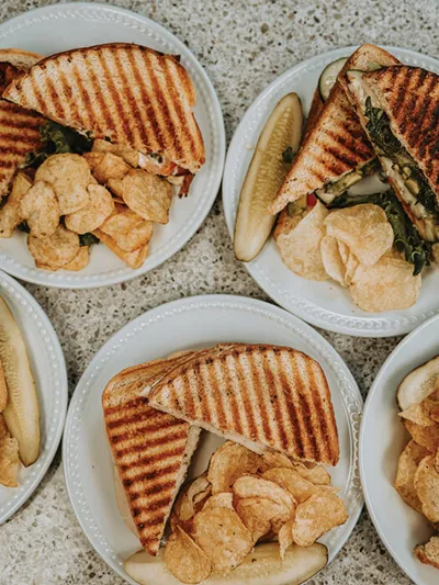 Four white plates with grilled panini halves and potato chips, one with a pickle spear