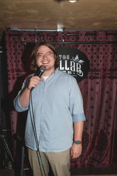 Smiling comedian holding a microphone on stage in front of The Cellar venue backdrop