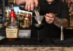 Tattooed bartender pouring Maker's Mark whiskey into a metal jigger