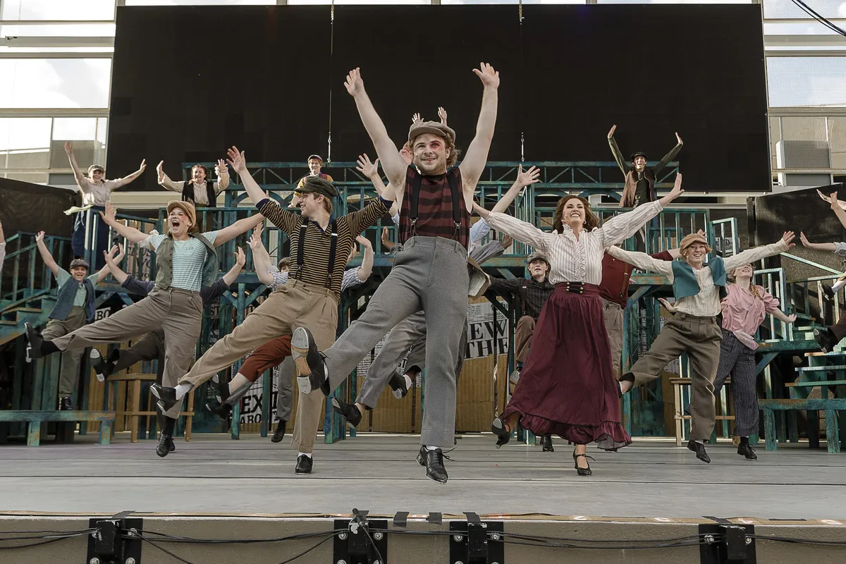 Large ensemble cast in period costumes leaping mid-air on an outdoor stage