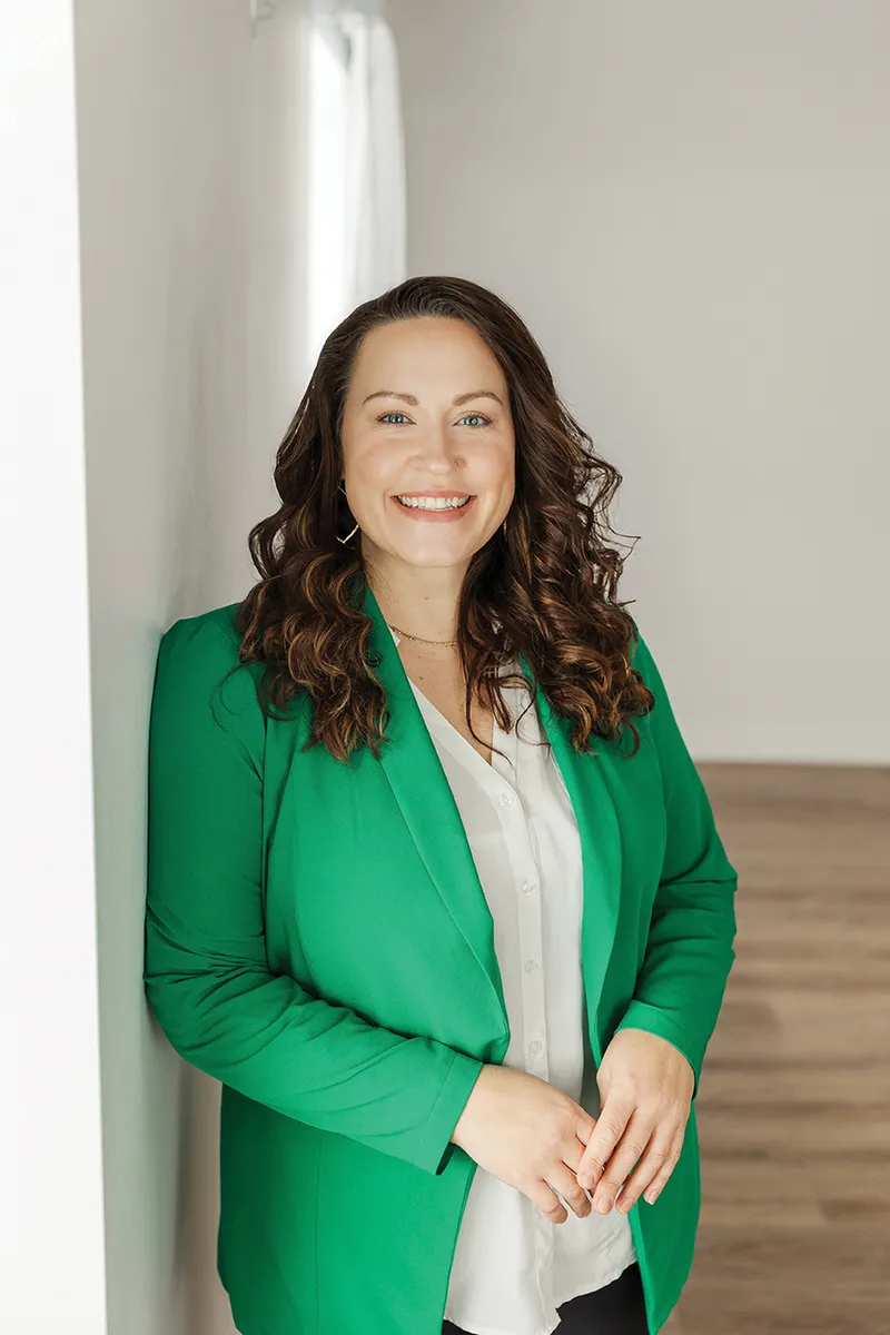 Smiling woman with curly brown hair wearing a green blazer and white blouse, leaning against a white wall