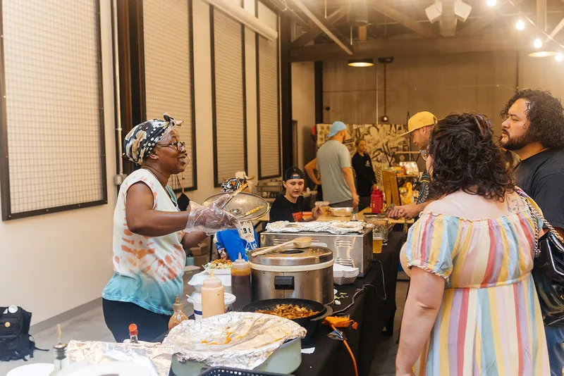 Woman in head wrap serves food at an indoor vendor table to waiting guests