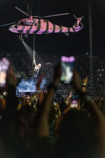 Performer rappels from a pink camouflage helicopter above a packed concert crowd at night
