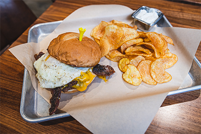 Burger topped with a fried egg and peppers beside kettle chips on a metal tray