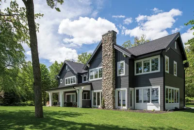 Dark gray two-story home with stone chimney, white trim, and manicured lawn surrounded by trees