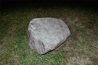 Large rough stone resting on grass at night, faintly inscribed