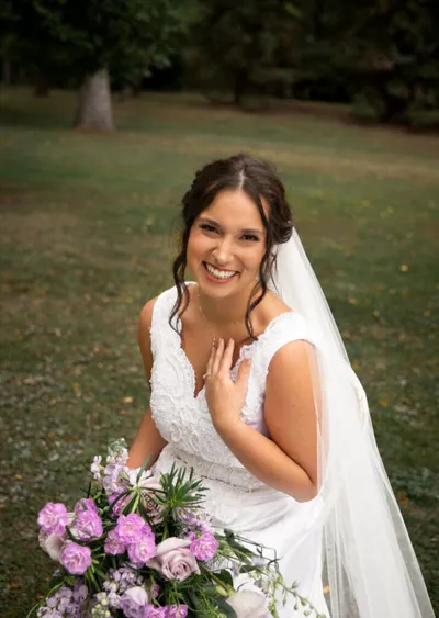 Smiling bride in white lace gown and veil holding a pink and lavender bouquet on a grassy lawn