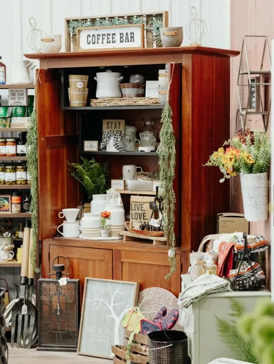 Wooden armoire styled as a coffee bar display with white ceramics, plants, and decorative signs in a shop