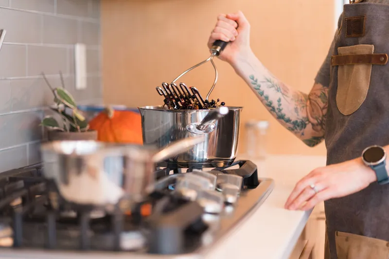 Tattooed person in brown apron mashing dark berries in a steel pot on a gas stove